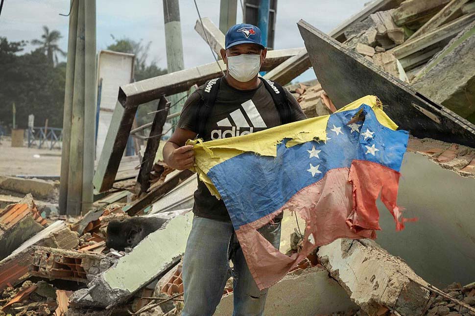 A Venezuelan citizen holds a national flag next to his damaged r
